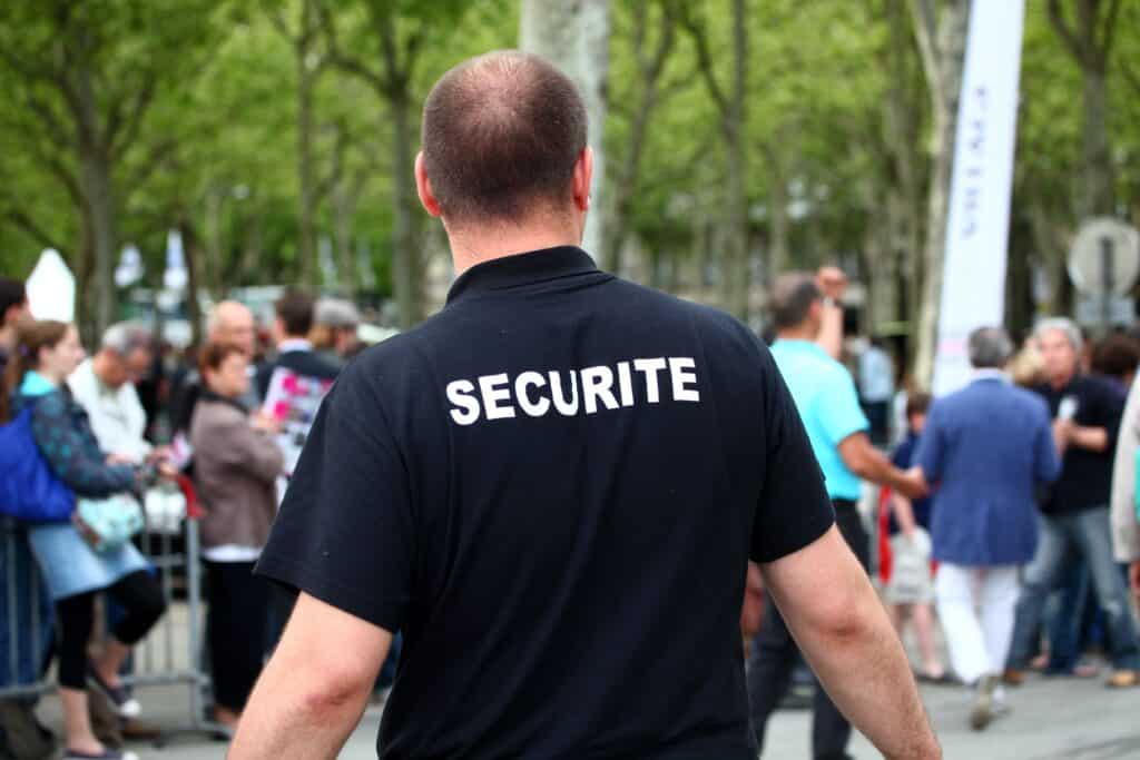 Personne vue de dos avec polo noir "SECURITE", parmi une foule en extérieur avec arbres et barrières.
